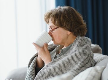 Ältere Dame mit Decke sitzt im Sessel und trinkt eine Tasse Tee